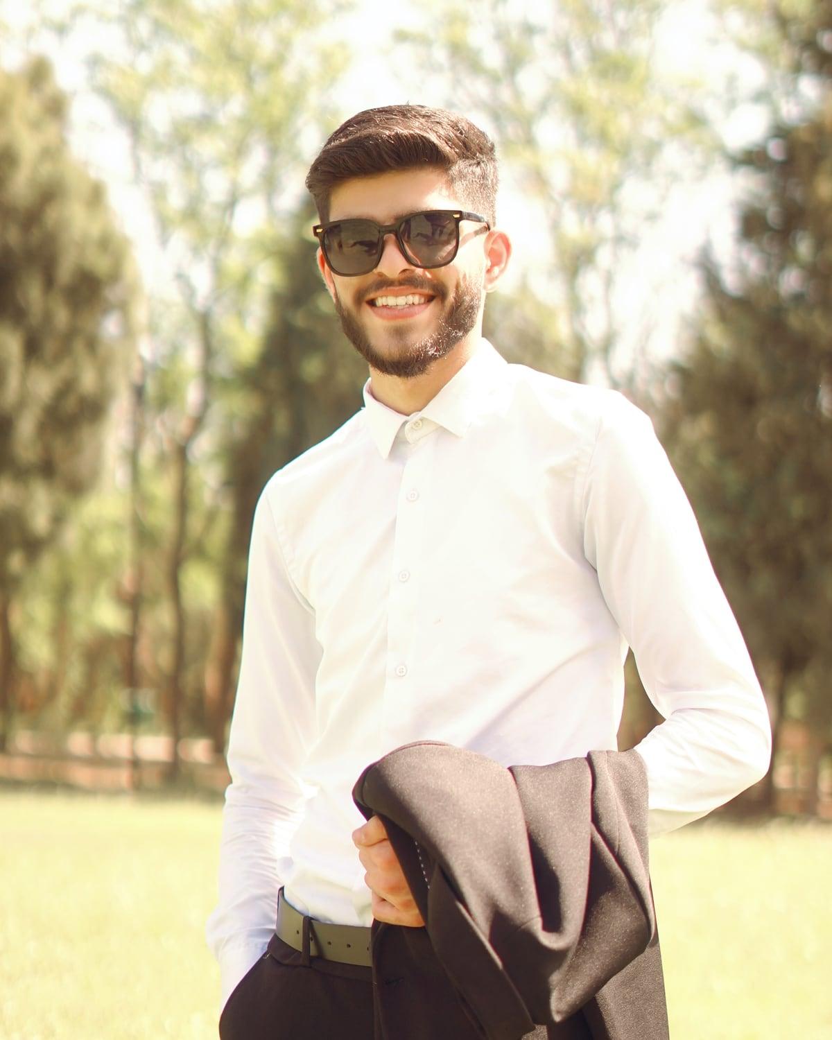 Ahmad Hiwa smiling in a white shirt and sunglasses, holding a dark jacket, in a park